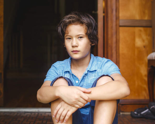 A boy with golden skin and serious expression sits in doorway of home — Stock Photo