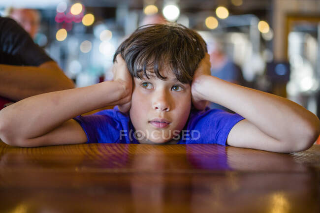 Un chico con la cabeza en las manos se sienta en la mesa del restaurante esperando pacientemente - foto de stock