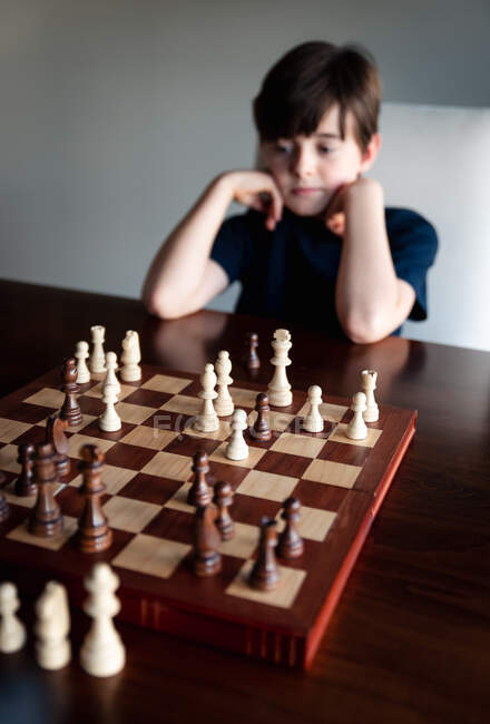 Молодой задумчивый мальчик сидит за шахматной доской и смотрит на фигуры. — стоковое фото