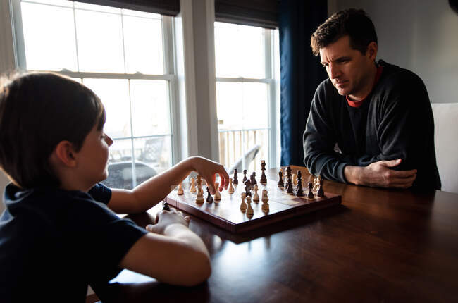 Padre e hijo sentados en una mesa adentro jugando un juego de ajedrez. - foto de stock