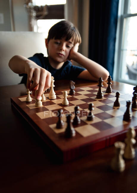 Joven chico pensativo sentado detrás del tablero de ajedrez moviendo una de las piezas. - foto de stock