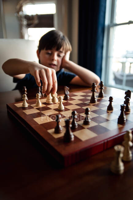 Jovem menino pensativo sentado atrás do tabuleiro de xadrez movendo uma das peças. — Fotografia de Stock