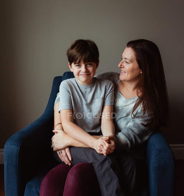 Happy woman hugging her son as he sits in her lap on a chair. — Stock Photo