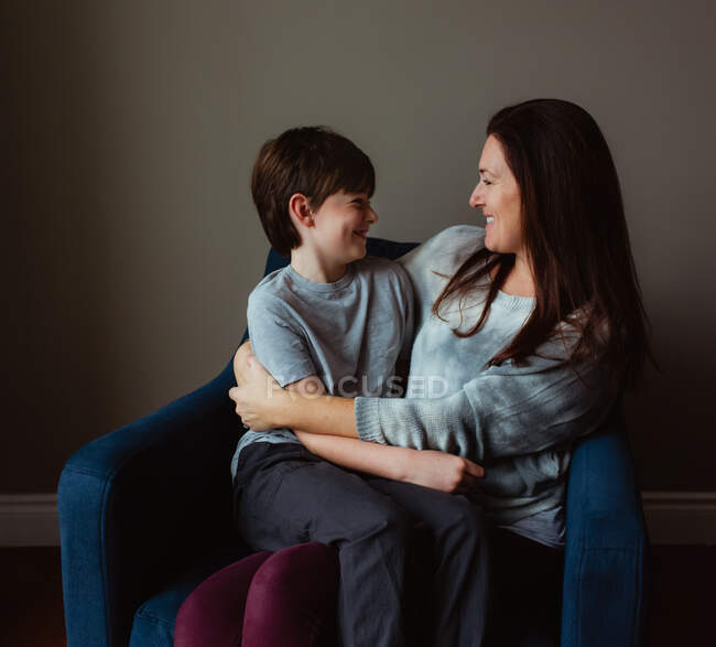Glückliche Frau umarmt ihren lächelnden Sohn, als er auf ihrem Schoß auf einem Stuhl sitzt. — Stockfoto