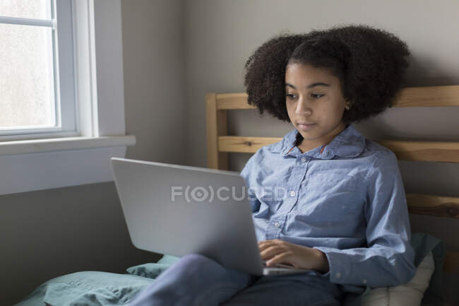 Niña bi-racial de once años que trabaja en la computadora portátil en la cama - foto de stock
