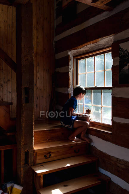 Niño sentado en las escaleras de cabaña cabaña de madera mirando por la ventana - foto de stock