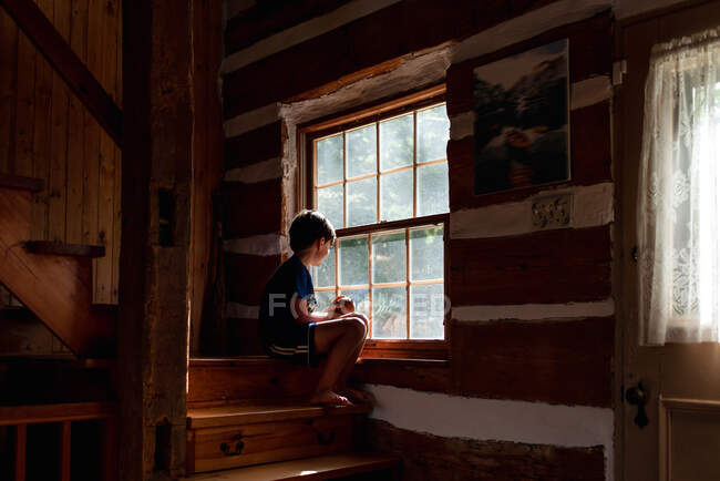 Garçon assis sur les marches d'un chalet en rondins regardant par la fenêtre — Photo de stock