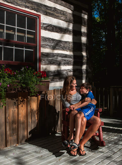 Mãe e filho sentados em cadeira de balanço no convés de uma cabine rústica. — Fotografia de Stock