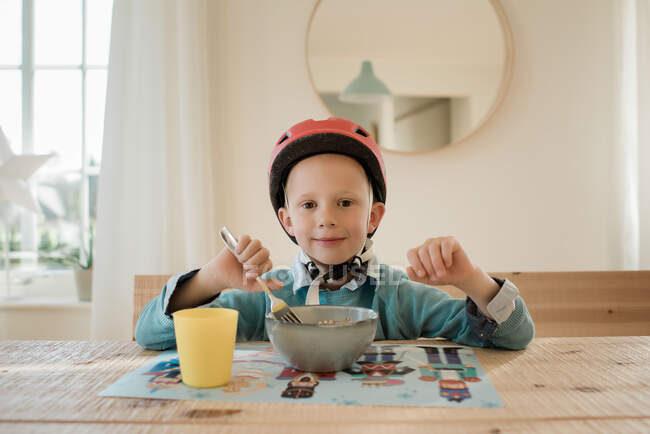 Niño se sentó en la mesa de la cena sonriendo con su casco en ansioso de ciclo - foto de stock