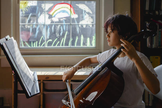 Сфокусований хлопчик сидить перед пофарбованим вікном, що практикує віолончель — стокове фото