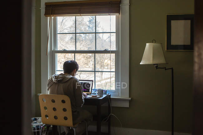 Un hombre se sienta en el escritorio frente a la ventana en el dormitorio trabajando en un ordenador - foto de stock