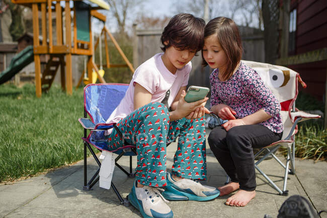 Dos niños se sientan con la cabeza junta mirando el dispositivo móvil - foto de stock
