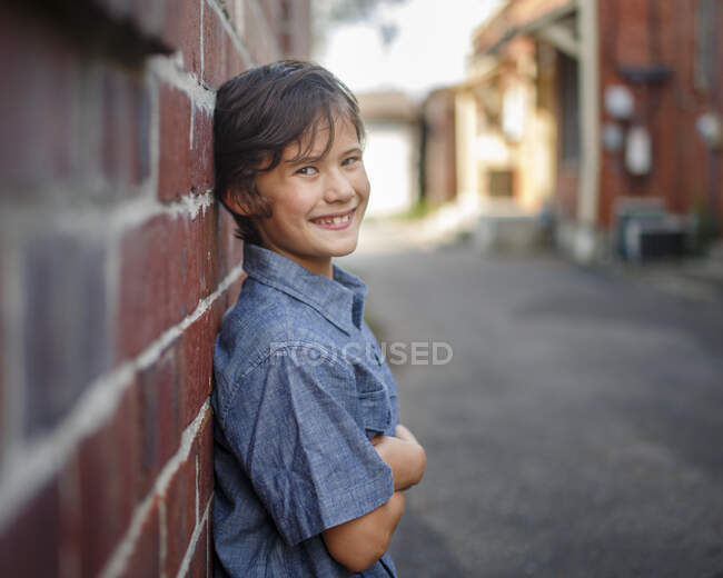 Un niño sonriente de pie en un callejón iluminado por el sol se apoya contra la pared de ladrillo - foto de stock