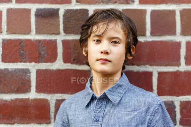 Un chico serio y hermoso se apoya en la pared de ladrillo con una mirada directa - foto de stock