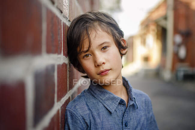 A beautiful serious boy leans against wall in sunlit brick alley — Stock Photo