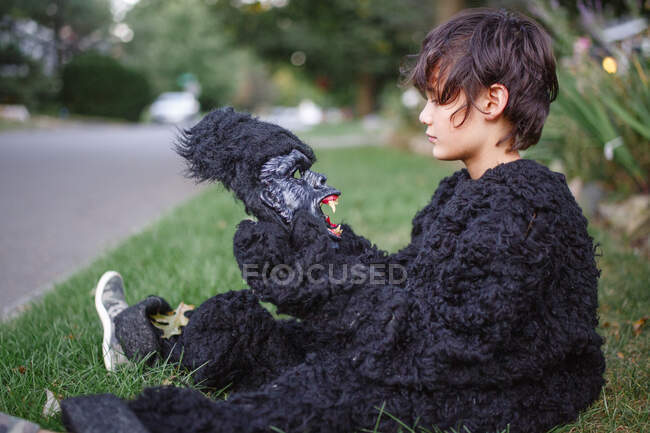 Un ragazzo siede in erba in tuta gorilla guardando spaventosa maschera gorilla — Foto stock