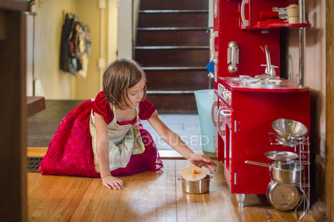 Маленька дівчинка в фартусі і вечірньому платті грає з іграшковою кухнею — стокове фото