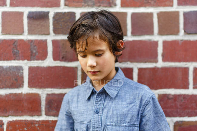 Portrait of a young boy leaning against brick wall shyly looking down — Stock Photo