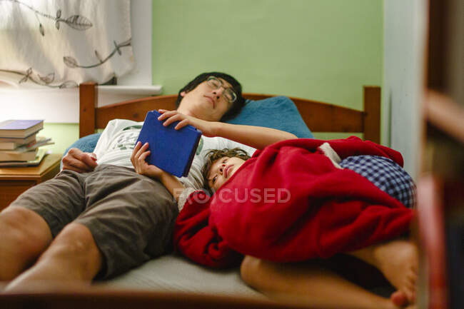 A boy cuddles up to his sleeping father in bed reading book at bedtime — Stock Photo