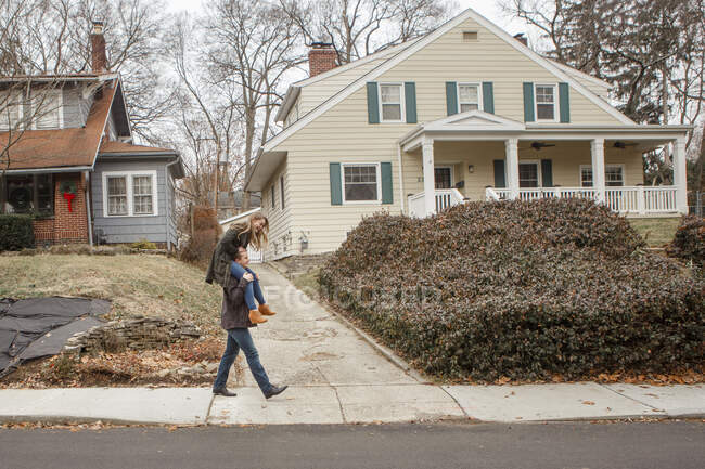 A young man carries his sister piggy back down a suburban sidewalk — Stock Photo