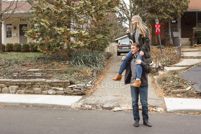 Un hermano adolescente lleva a su hermana riéndose sobre sus hombros en la calle - foto de stock