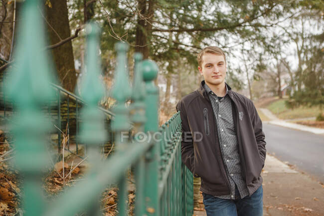 A serious young man leans against a wrought iron fence  in autumn — Stock Photo