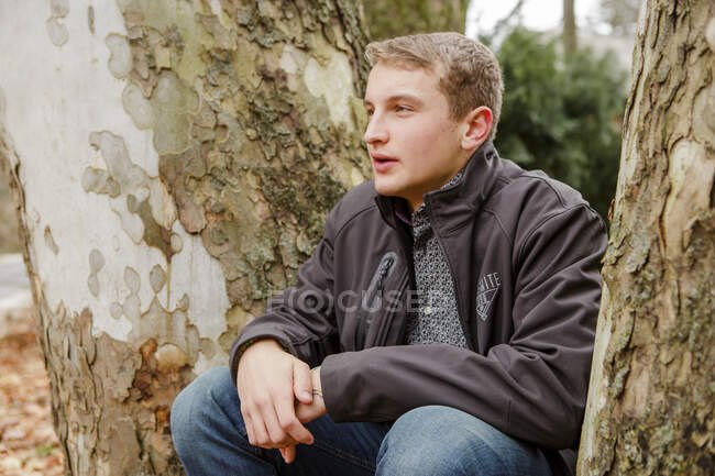 Un adolescente se sienta afuera apoyado contra un sicómoro en otoño - foto de stock