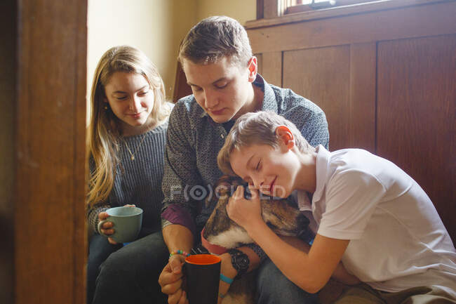 Trois frères et sœurs heureux câlinent leur chien dans une belle fenêtre dorée — Photo de stock