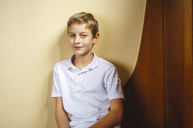 Un jeune garçon s'assoit sur un banc en bois appuyé contre un mur avec un regard direct — Photo de stock
