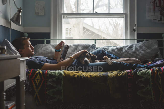 Un joven se acuesta en la cama acariciando al perro en su regazo y leyendo un libro - foto de stock