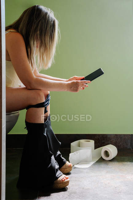 Блондинка на туалеті дивиться телефон, поки вона пісяє або чхає. Вертикальне фото — стокове фото