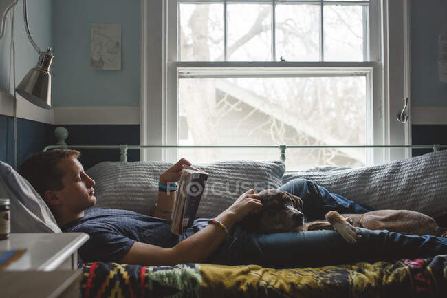 A young man lays on his bed with dog on lap reading by window light — Stock Photo