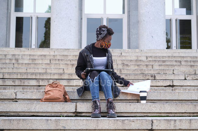 University female african student wearing protective face mask studying sitting on stairs outside on campus. New normal in college. — Stock Photo