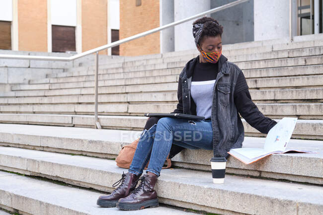 Studentessa africana universitaria con maschera protettiva che studia seduta sulle scale all'esterno del campus. Nuovo normale al college. — Foto stock