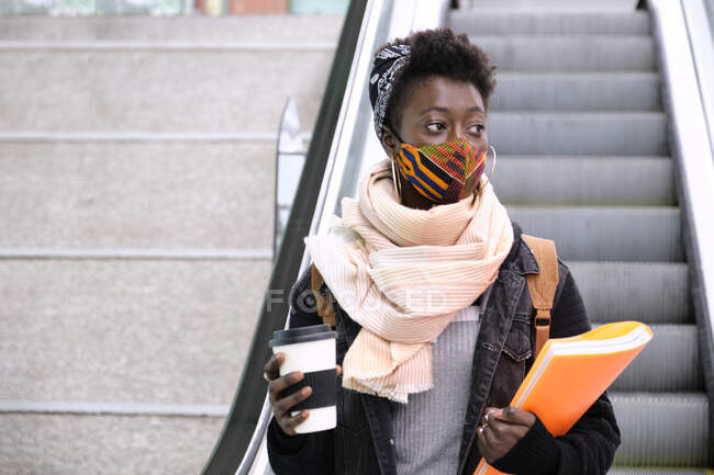 Étudiante africaine de l'université portant un masque protecteur prenant un escalator à l'étage de la station de métro. Nouveau normal dans les transports publics. — Photo de stock