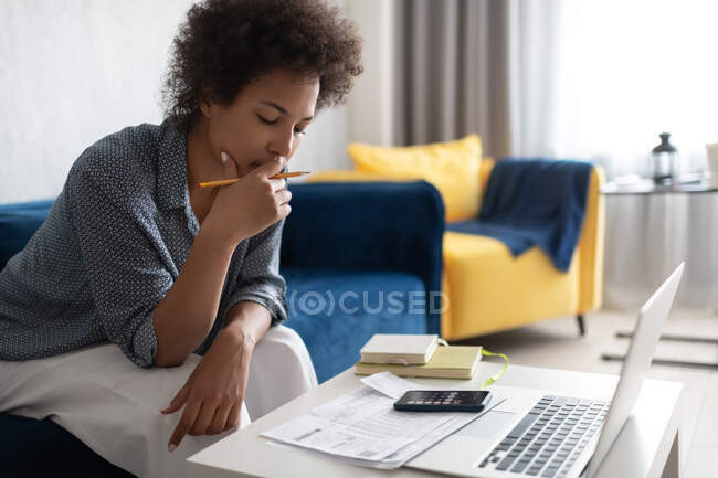 Preto feminino tocando rosto e pensamento enquanto sentado no sofá e pagar impostos — Fotografia de Stock