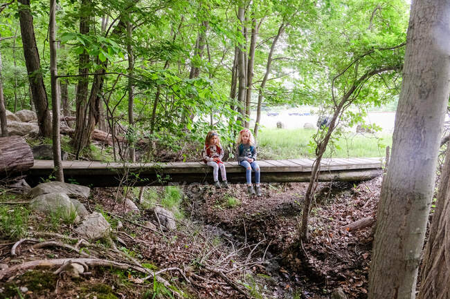 Dos chicas sentadas juntas y descansando en el puente en el bosque mientras caminan - foto de stock