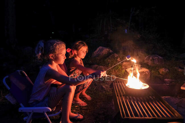 Two girls roasting marshmallows over open fire, camping in summertime — Stock Photo