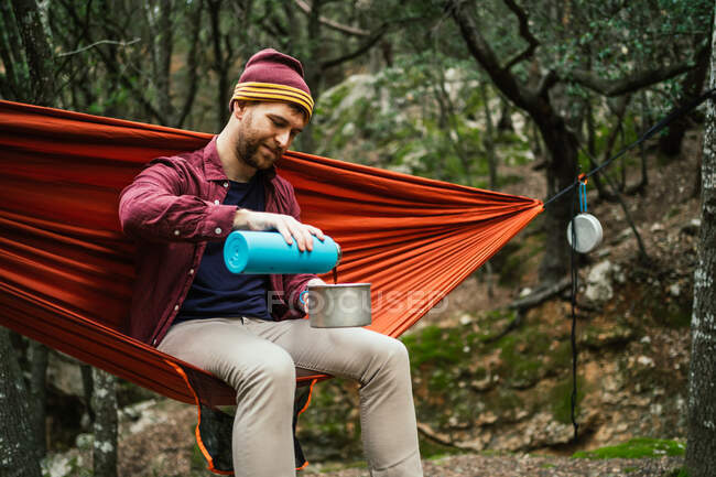 Человек наливает горячий напиток на горшок, сидя на гамаке в лесу — стоковое фото