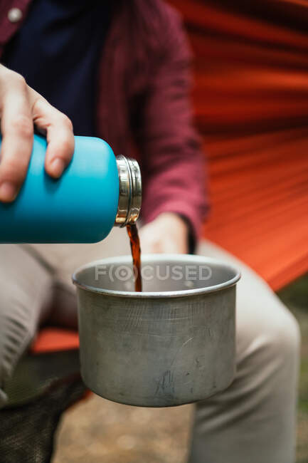 Homem derrama bebida quente em um pote sentado em uma rede na floresta — Fotografia de Stock