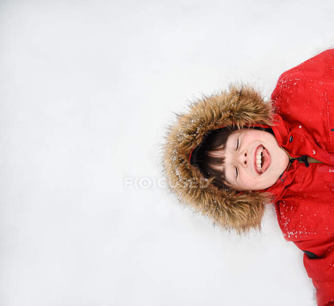 Накладка щасливого хлопчика в червоному пальто з пухнастим капюшоном, що лежить на снігу . — стокове фото
