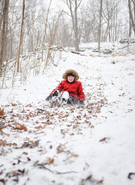 Rapaz feliz a descer uma colina na floresta num dia de inverno nevado. — Fotografia de Stock