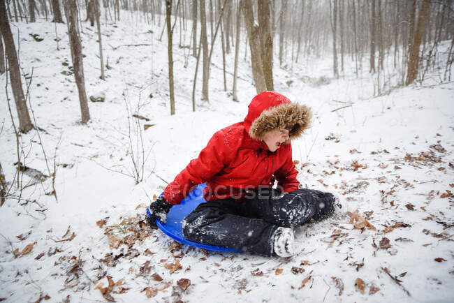 Felice ragazzo slittino giù per una collina nel bosco in una giornata invernale nevosa. — Foto stock