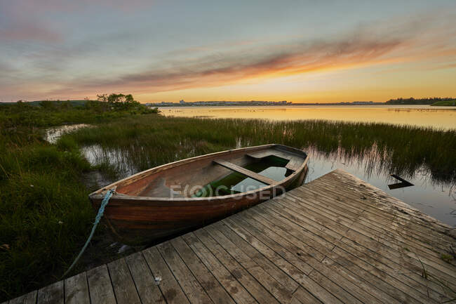 Поврежденная деревянная лодка с водой и деревянным пирсом, расположенная рядом с травянистым берегом и спокойным озером на фоне облачного заката — стоковое фото