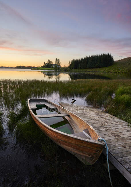 Barco roto y muelle de madera situado en el agua herbácea pacífica del lago contra el cielo del atardecer en verano en la naturaleza - foto de stock