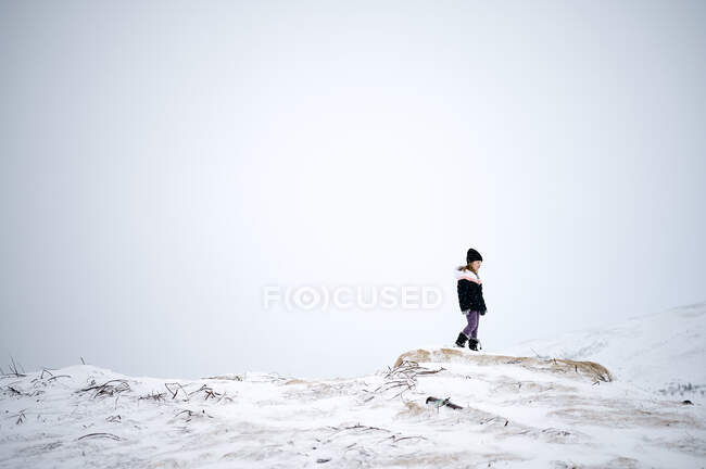 Вид сбоку стильной маленькой девочки в теплой верхней одежде и шляпе, идущей по раскаленной террасе, покрытой снегом, в солнечный зимний день — стоковое фото