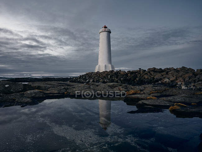 Torre do farol localizado em penhasco rochoso perto de água do mar reflexiva calma contra o céu nublado cinza — Fotografia de Stock