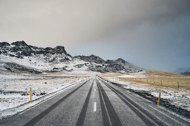 Estrada de asfalto em linha reta que vai em direção ao cume da montanha coberto de neve contra o céu nublado na natureza — Fotografia de Stock