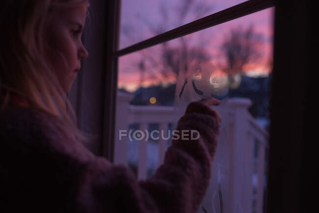 Kleines Mädchen schreibt auf Fensterkondenswasser mit Sonnenuntergang im Hintergrund — Stockfoto