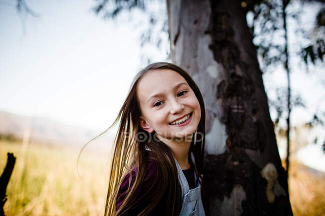 Chica joven sonriendo para la cámara al lado del árbol - foto de stock